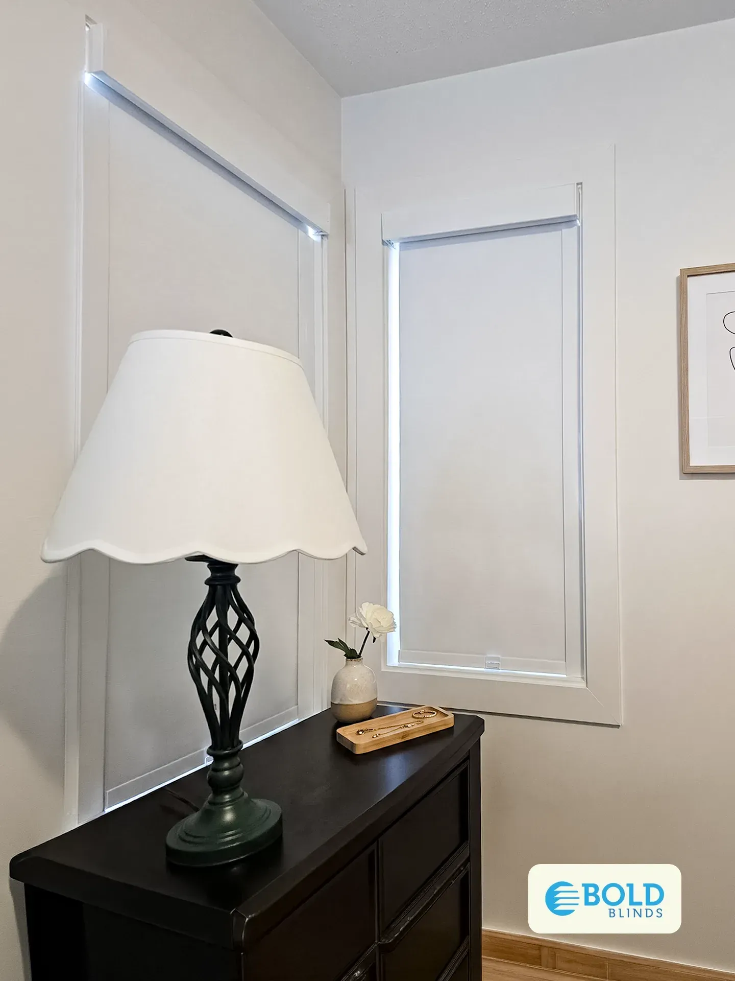 Lowered white roller shade on a bedroom window beside a lamp and dresser