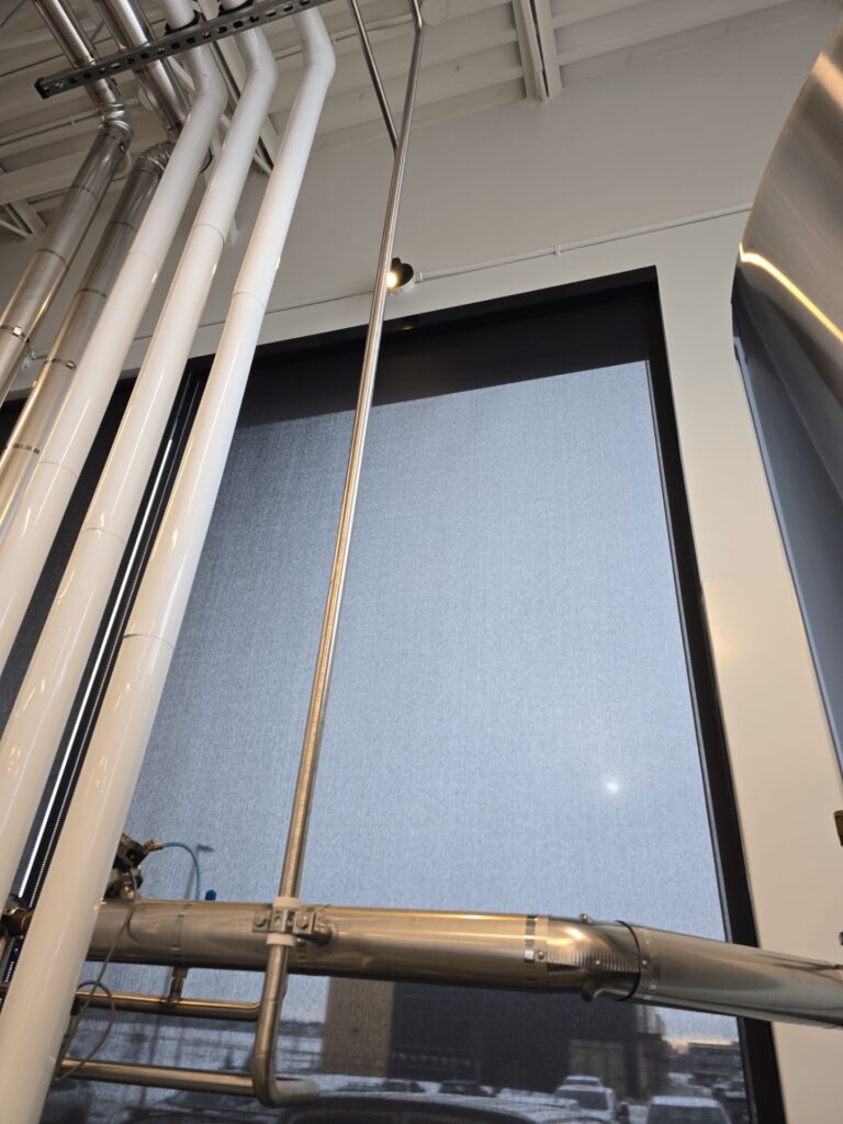 Solar roller shades (3–5% openness) cutting glare in an Edmonton taproom while preserving views of stainless tanks.